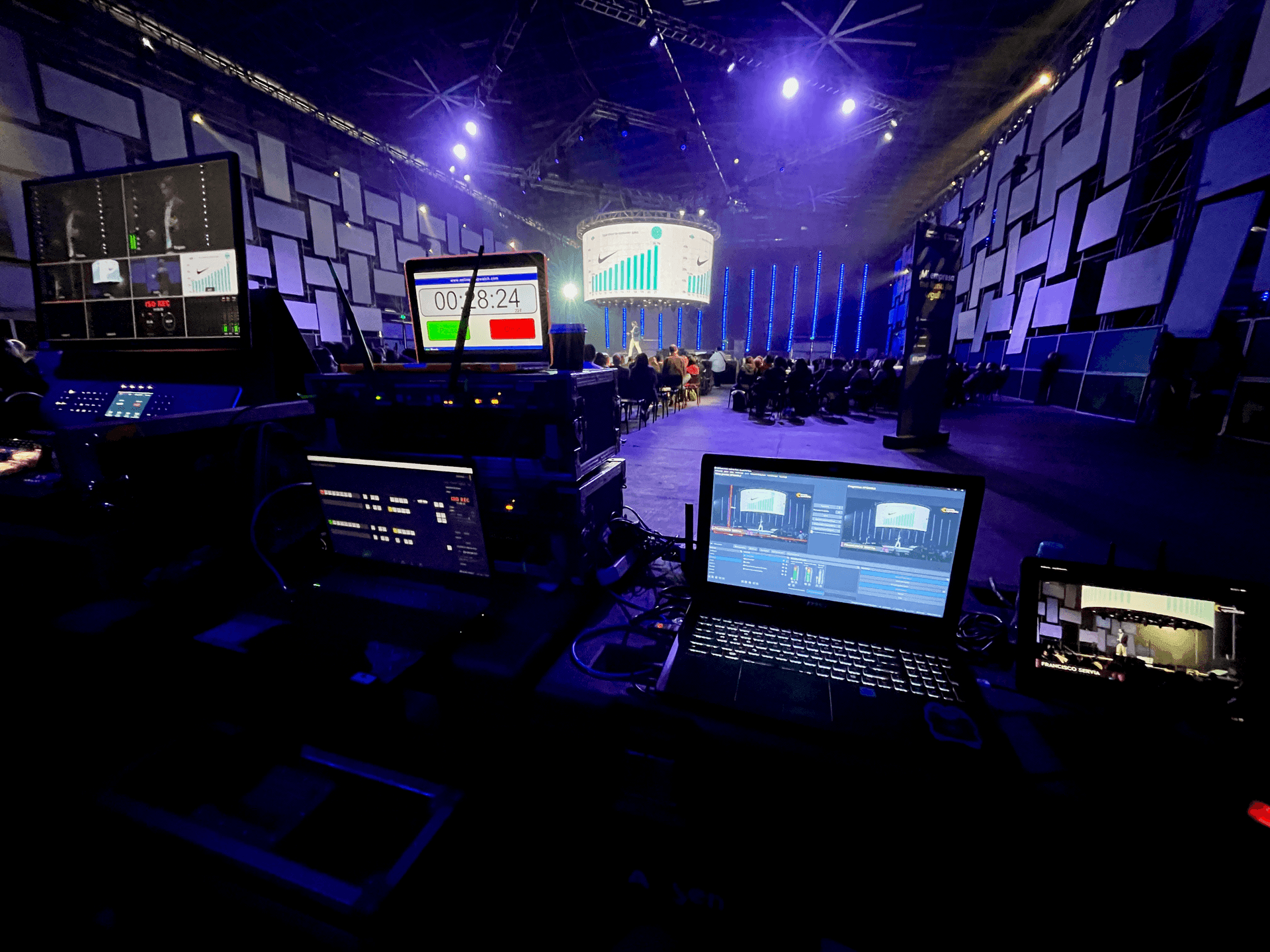 Backstage view of a conference with computers and screens displaying live feed and countdown timer.