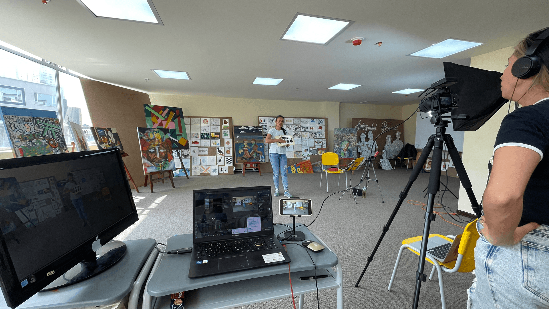 Art exhibition setup with paintings, camera on tripod, and person standing in the background holding artwork.