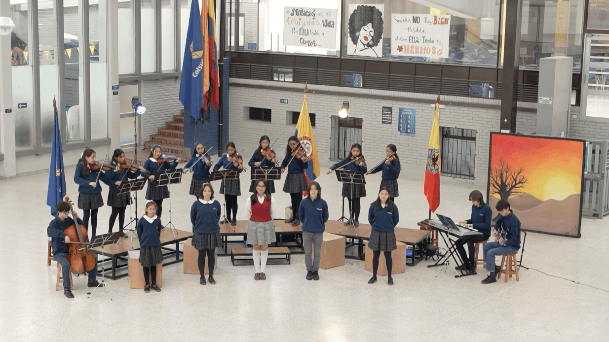 School orchestra performance with students playing string instruments, keyboard, and guitar in a hall.