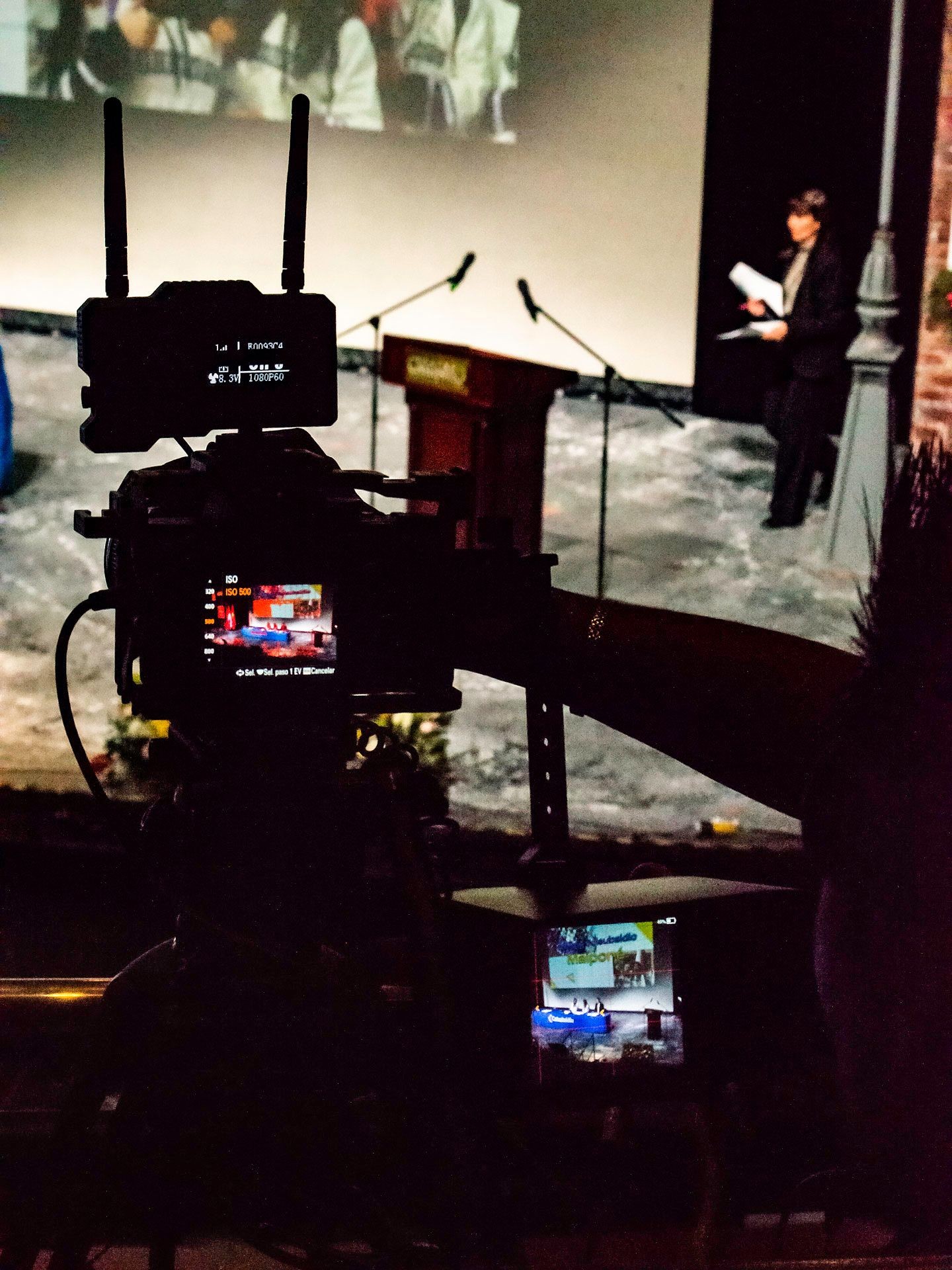 Camera filming a stage with podium and microphones, person holding papers in the background.